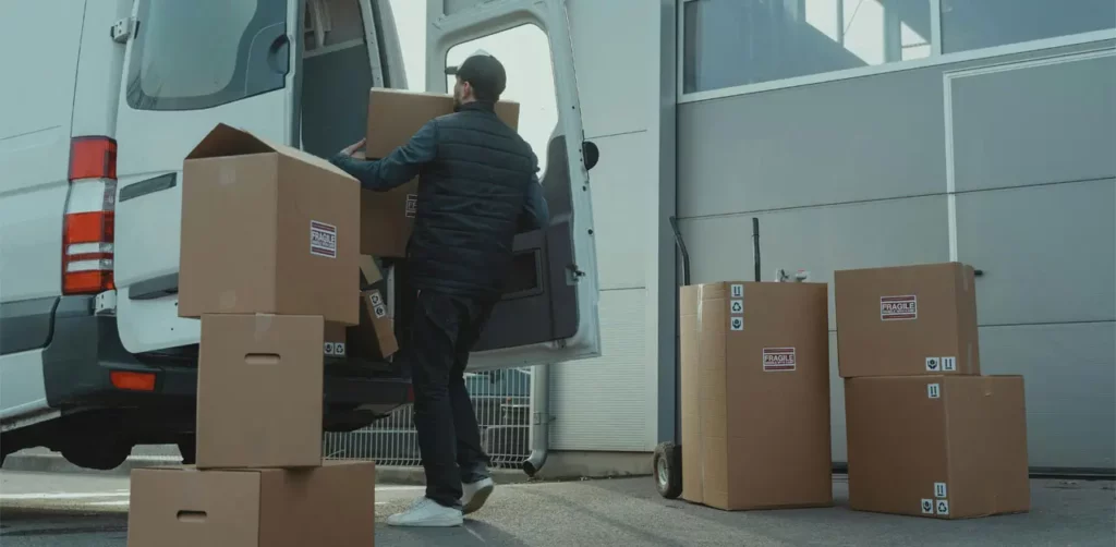 A Man Is Loading Cardboard Boxes Into The Back Of A Van, Preparing For Transportation