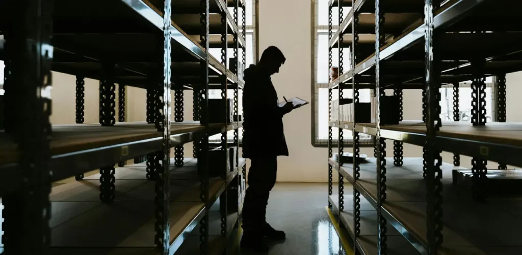 A Person Stands In A Warehouse Surrounded By Tall Shelves Filled With Various Items, Highlighting The Storage Environment